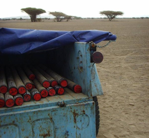 Light blue open-backed truck with drilling cores. African arid landscape with sparse trees in the background.