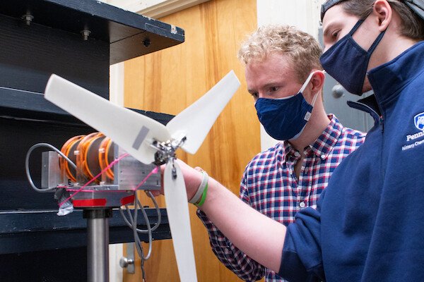 Two young men wearing masks adjust a mini-propellor, a portion of a model wind turbine. 