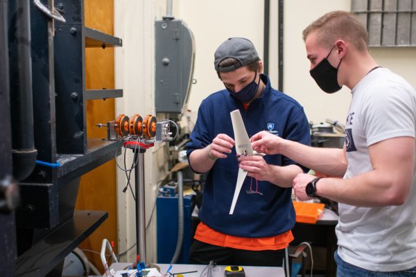 Two young men wearing face masks assemble a small wind turbine, which includes a motor, several cords and a white-winged propeller. 