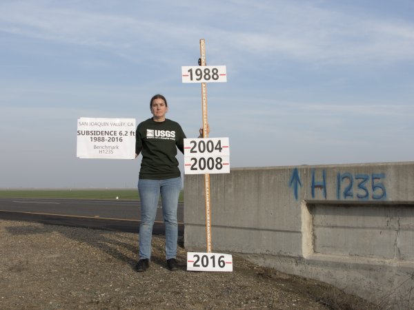 USGS scientist standing with a pole annotated with land-surface elevation marks at given years at bench mark H 1235 RESET.