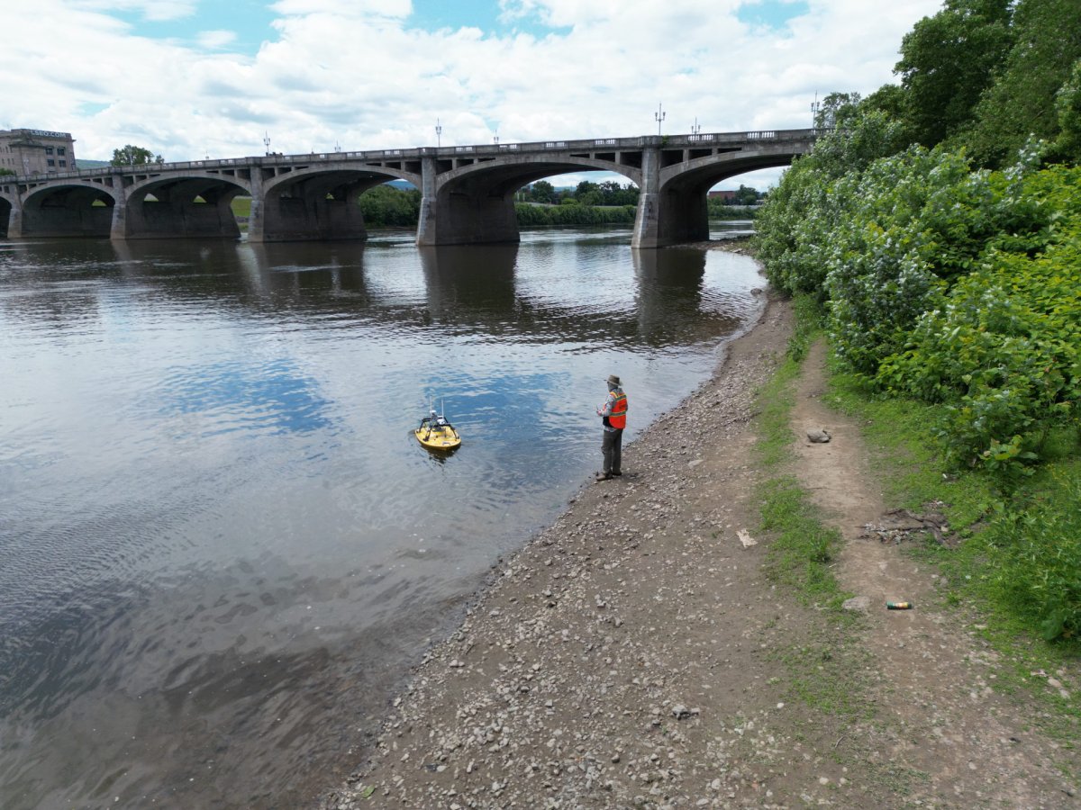 An operator stands on the shore of a river near a large bridge, operating a remote controlled boat a few feet from shore.