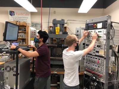 two students in an engineering lab, one student is on a computer and the other student is turning dials on a large machine